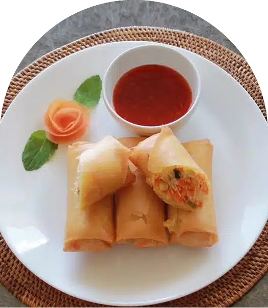 Four golden spring rolls are arranged on a white plate with a small bowl of red dipping sauce. A carved carrot flower and two mint leaves decorate the side. One spring roll is cut open, showing its vegetable filling.