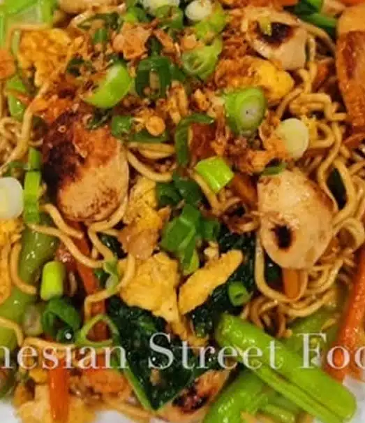 A close-up of Indonesian street food featuring stir-fried noodles with chicken, vegetables, scrambled eggs, and topped with chopped green onions. The dish looks colorful and appetizing.